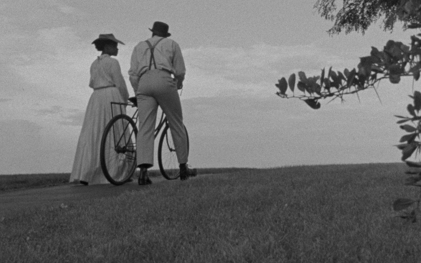 A woman in a long dress and a man walk together with a bicycle away from the camera over a grassy hill.