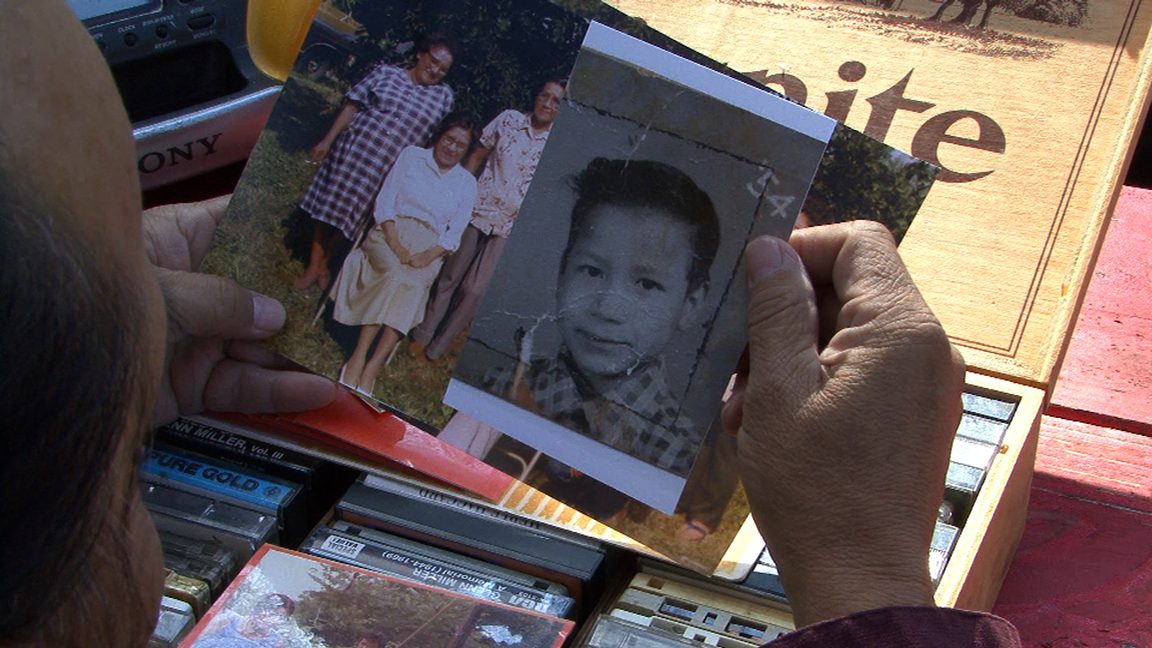 An over-the-shoulder shot of a person holding family photos over a box of casettes, one on top of a young boy’s school photo.