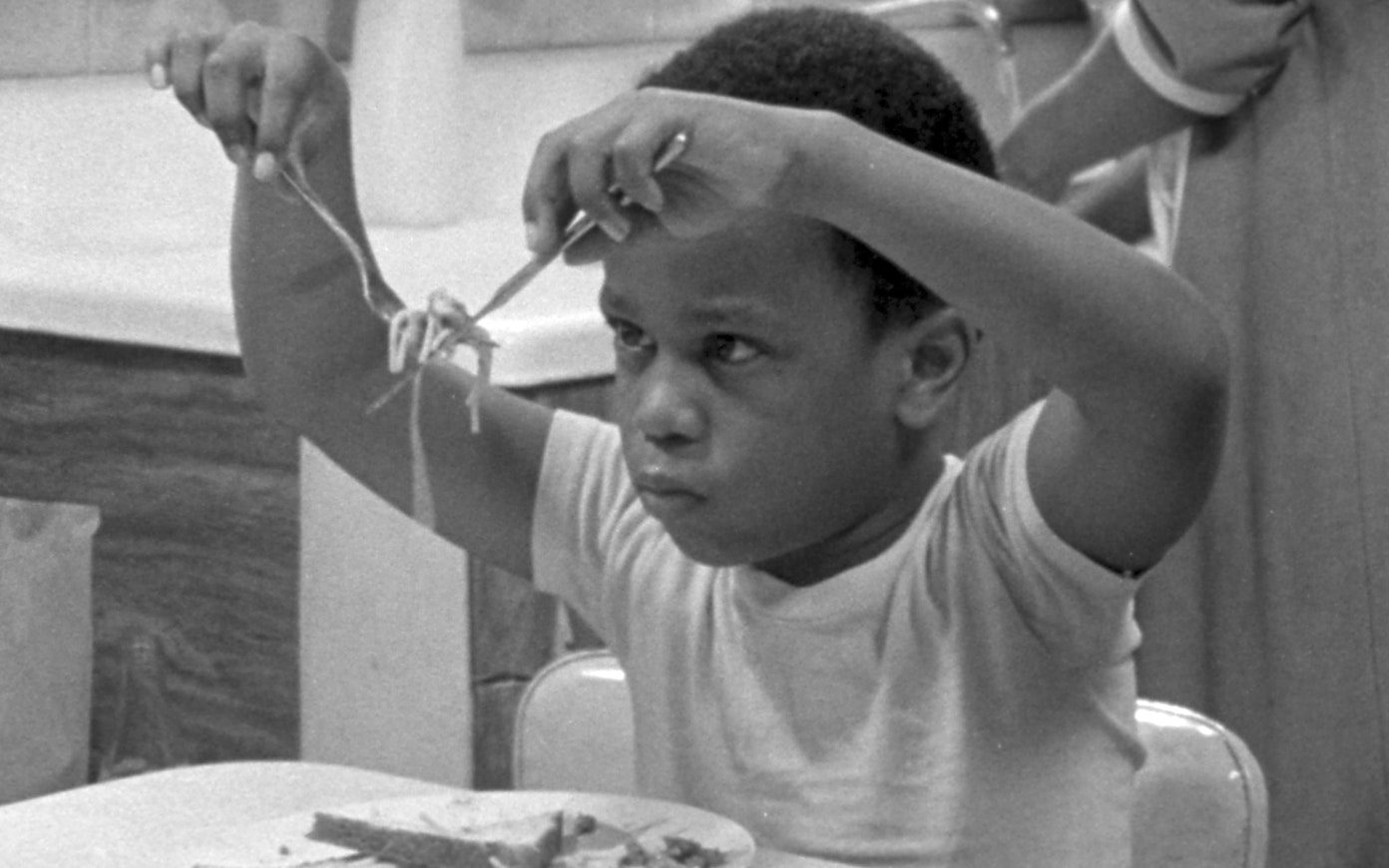 Black and white still of a little boy sitting at a dining table holding up noodles with a fork and knife in front of his face and staring at it.