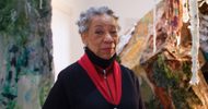 An older woman with short gray hair and a warm smile stands in an art studio, wearing a red top, black cardigan with white patterns, and gray pants. Colorful, textured art pieces and materials surround her.