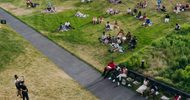 People are sitting on grass and benches in small groups at an outdoor event, spaced apart on a green lawn, with a few standing near a paved path. The scene is relaxed and summery.
