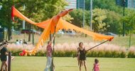 Several people hold up a large, bright orange bird puppet on poles in a sunny park, while a small child in a red dress stands nearby. Tall buildings and green trees are visible in the background.