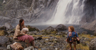 On a rocky shore beneath a waterfall, a man with light skin sits and holds a baby by another man with light skin who has dwarfism and curly red hair.