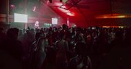 A large crowd of people is dancing and socializing inside a dimly lit venue with red lighting. Two bright screens and a performer are visible on a stage in the background.