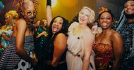 A group of people dressed in colorful, festive outfits smile and pose together at a lively party with bright lights and a joyful atmosphere.