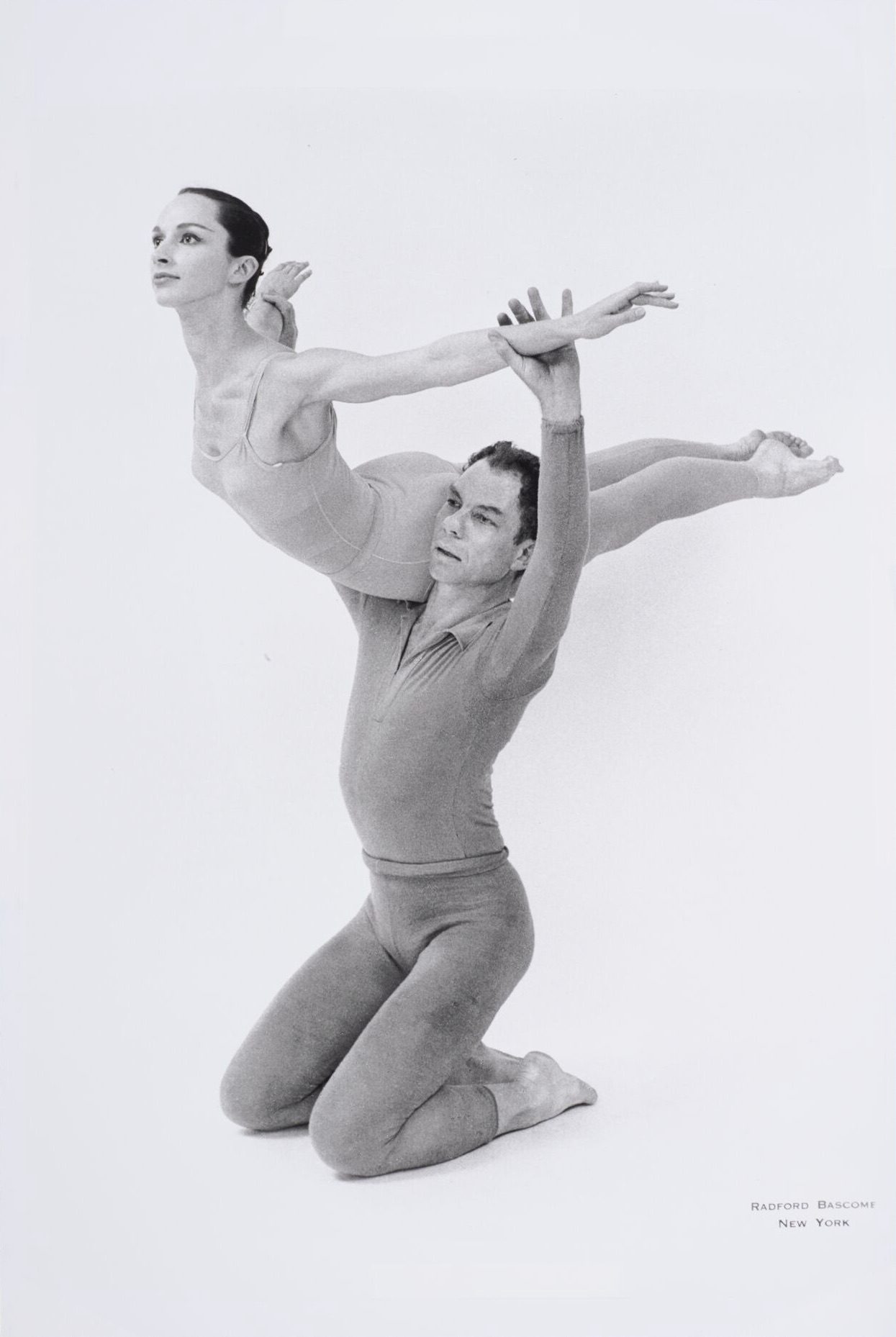 Two ballet dancers perform a lift; one dancer is kneeling while supporting the other, who is extended horizontally with arms outstretched, both wearing fitted dance attire against a plain background.