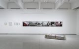 Art gallery with framed photos on a white wall and a wrapped object lying on the floor in front.