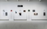 Artworks and framed texts arranged on a white gallery wall with two large text panels leaning against the wall.