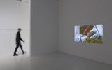 A person walks past a white gallery wall where a video of an elderly person lying in bed is projected. The space is minimalistic and well-lit, with a smooth, empty floor.