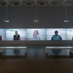 A gallery room with five large video projections showing people standing by the sea, their backs or profiles facing the camera. Two white benches sit in front of the screens on the polished floor.