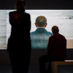Two people are silhouetted in front of a large screen displaying a video of a person in a denim jacket sitting on a beach and facing the ocean.
