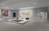 A large gallery space with objects in the middle and paintings on a grey wall.