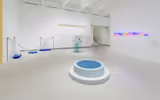 A white gallery space with a blue pool like sculpture in teh middle and some other sculptures around.