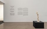 A small white sculpture of a standing nude figure is displayed on a black pedestal in a gallery. Text on the wall includes “Sculpture Court” in multiple languages and exhibition information. The space is minimal and well-lit.