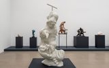 A modern art gallery features a large abstract white sculpture in the foreground, surrounded by various colorful and dark-toned figurative sculptures displayed on pedestals in the background against a white wall.