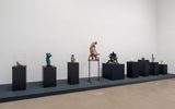 A row of eight various abstract sculptures displayed on black pedestals, some covered by glass cases, in a minimalist gallery with white walls and a smooth concrete floor.