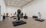 A spacious art gallery with white walls and high ceilings displays various modern sculptures on black platforms, including abstract forms, human figures, and organic shapes, all arranged across the open floor.