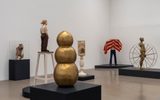 A contemporary art gallery displays various sculptures, including a gold three-sphere stack, a headless figure in a red-striped shirt, a doll, a taxidermy dog, and a human figure with a large metal wheel.