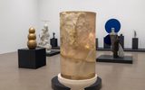 A modern art gallery displays various sculptures, including a large translucent cylindrical sculpture with two overlapping faces in the foreground, and abstract and figurative works on black platforms in the background.