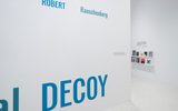 A minimalist white gallery wall features the large blue text ROBERT RAUSCHENBERG DECOY. In the background, shelves display small photo prints, and additional text is visible on another wall.