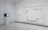A minimalist gallery space displays a large abstract artwork with black and blue lines on a white background and a small TV monitor on a pedestal showing a similar abstract image, against a backdrop of white walls and curtains.