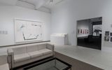 A modern art gallery with white walls, a large abstract painting, display panels, a glass coffee table, and cream sofas. An open doorway reveals another room with framed art and a spiral staircase.
