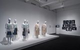 A museum exhibit displays several mannequins dressed in light-colored, modern garments. The room has white walls, and framed black-and-white photos hang on the wall in the background.