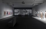 A modern gallery with art prints on the left wall, large grayscale abstract images on the back wall, and mannequins dressed in contemporary outfits displayed on a platform to the right. Minimal seating is in the center.