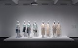 Eight mannequins dressed in light-colored, semi-transparent garments stand on a white platform in a minimalist gallery with a white wall and dark floor.