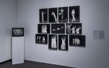 A museum wall displays black-and-white photos of dancers in various poses, arranged in a grid. To the left, a TV monitor on a pedestal shows a black-and-white video. Two information panels are mounted on the right.