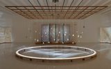 A large circular light installation on the floor of a gallery space is surrounded by suspended stones and a wooden grid overhead, with sheer curtains and subtle patterned wall panels in the background.