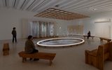 A modern art gallery with wooden benches, a large circular light installation in the center, hanging threads, and three people observing the exhibit in a minimalist, softly lit space.