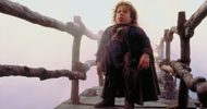 A person with dwarfism, light skin and wavy hair standing on a rustic bridge over a foggy white background wearing a blue shirt and a cape.