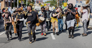 A diverse marching band plays instruments, including brass, drums, and saxophones, while walking down a city street during a daytime parade or protest. People hold signs in the background.