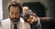 Three men with beards stand close together outside near a textured wall; one in a light suit looks downward, another in a dark suit rests his head on the first mans shoulder, and a third man stands behind them.