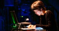 A person with short dark hair, wearing a black top, is focused on a laptop and electronic equipment on a dark stage, illuminated by blue lighting. Their motion creates a blurred effect on their face.