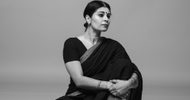 A woman in a black sari sits on the floor with her arms wrapped around her knees, looking thoughtfully to the side against a plain, light background. The photo is in black and white.