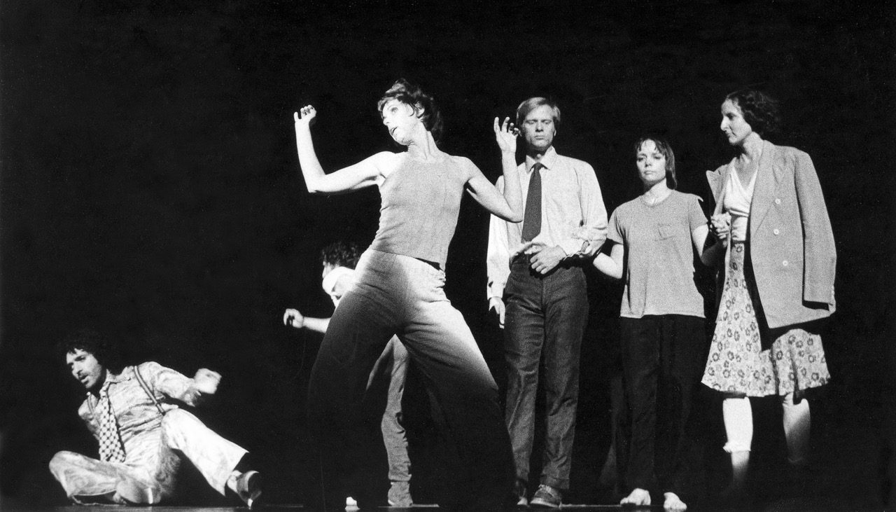 Black-and-white photograph of a theatrical performance. Six people are visible, including one central figure in an expressive stance and another seated or partially reclining on the floor at left. The other four stand nearby against a dark backdrop.