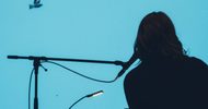 The back of musician Ishibashi seated at a laptop and mic-stand. She is silhouetted against a movie screen that is mostly turquoise with a flying bird.