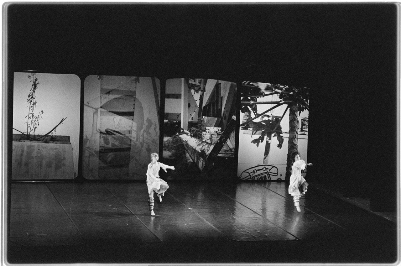 Two dancers in white costumes perform on a dark stage, in front of four large panels displaying black-and-white, abstract images of urban and natural scenes.