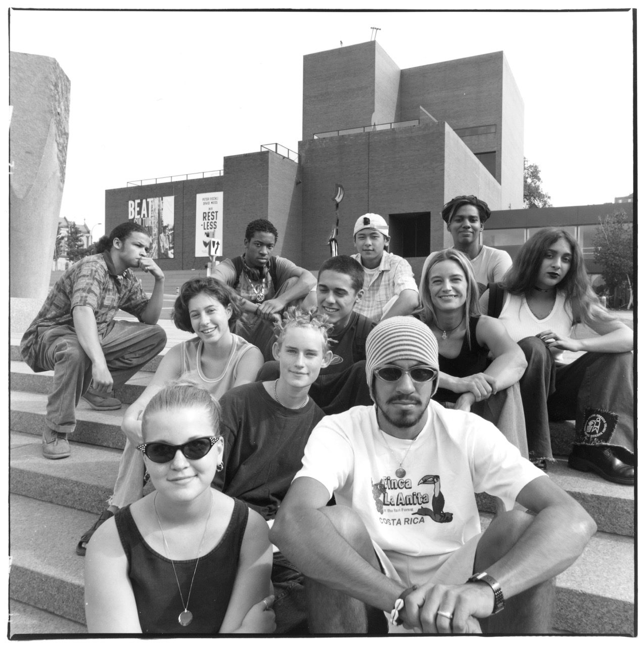 A diverse group of young adults sits together on outdoor steps, smiling and relaxed, with a modern brick building (the Walker Art Center) in the background.