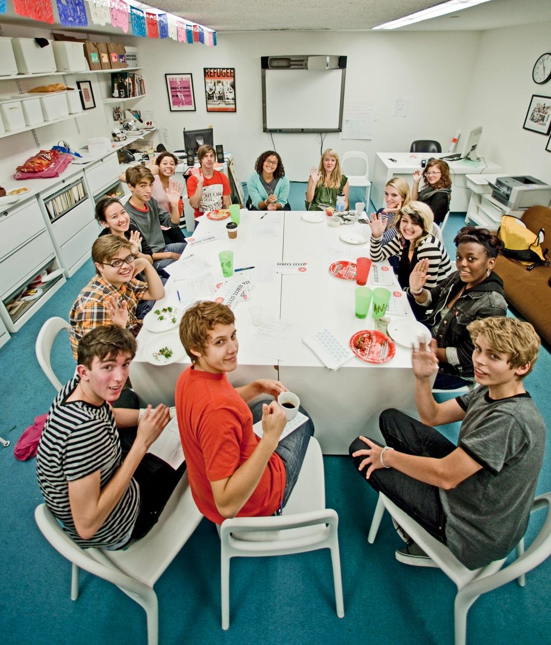 A group of teenagers sit around a large square table in a classroom, smiling and waving at the camera while eating snacks. Papers, cups, and plates are on the table. Bright decorations hang above.