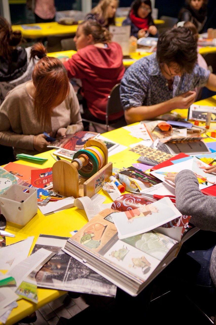 People sit around a yellow table covered with art supplies, books, and papers, working on various crafting projects in a brightly lit, busy room.