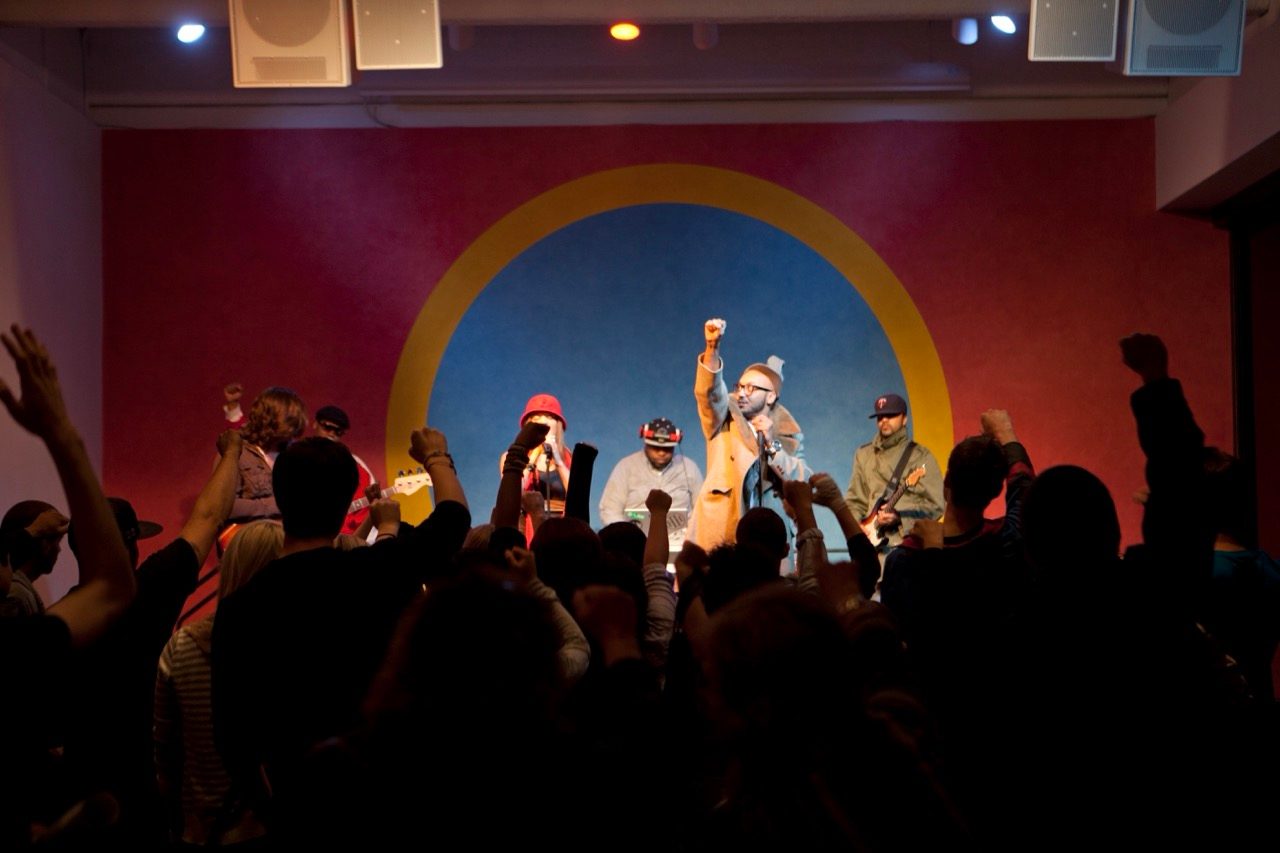 A band performs on stage with colorful background as the audience raises their fists in the air, showing solidarity and enthusiasm.