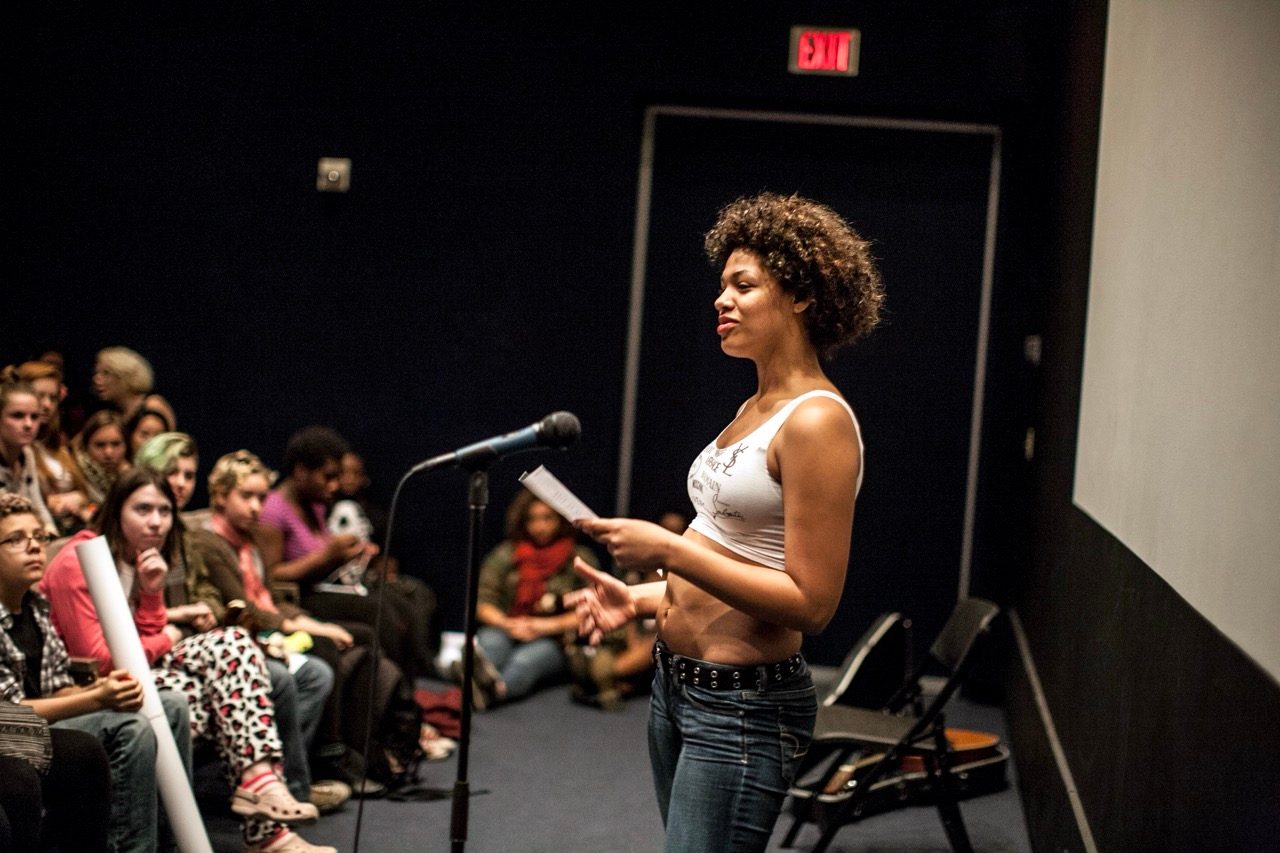 A woman stands at a microphone, speaking to a seated audience of young people in a dimly lit room. She holds papers in one hand.
