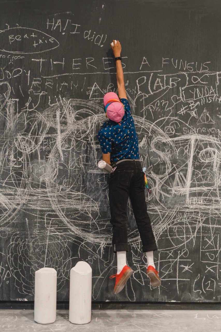 A person in a pink cap and blue shirt leaps in the air, reaching up to write on a large chalkboard covered with doodles and messages. Two white cylindrical oversized stubs of chalk are in front of the board.