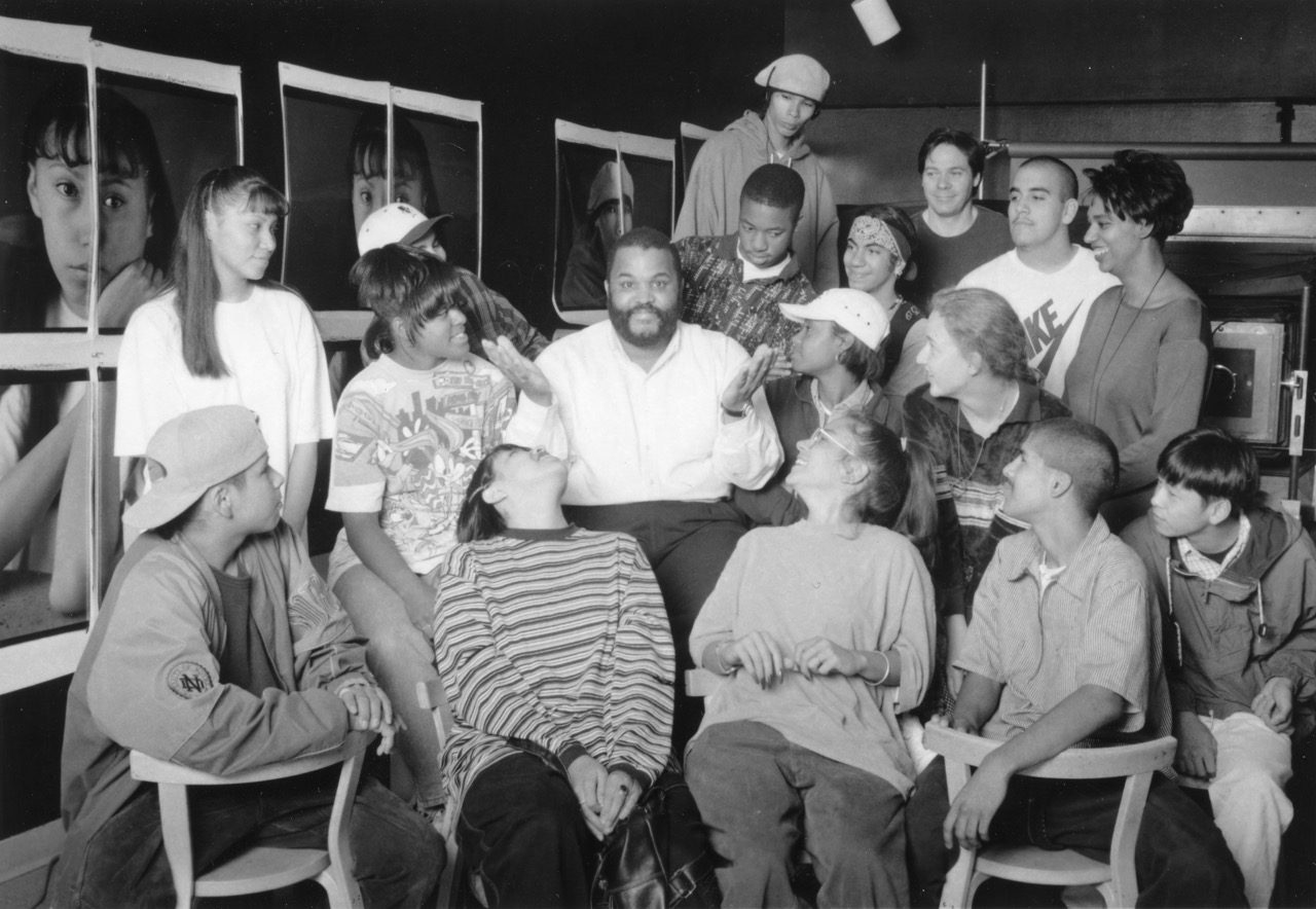 A group of teens sit and stand around an adult man in a white shirt, all engaged in conversation and laughter. Framed black and white photos decorate the background.