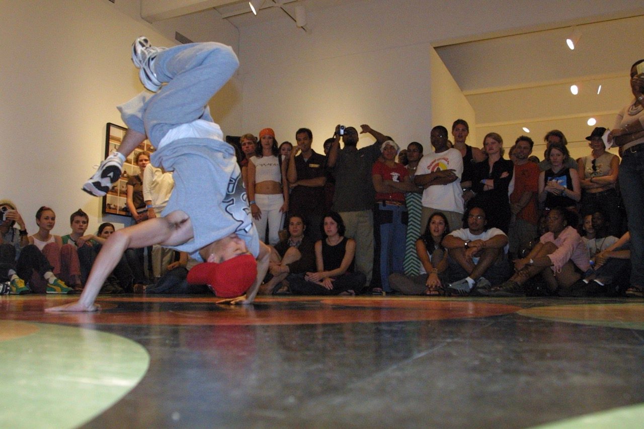 A breakdancer balances on their hands with legs in the air, performing on a colorful floor. A large, diverse crowd sits and stands around, watching and taking photos.