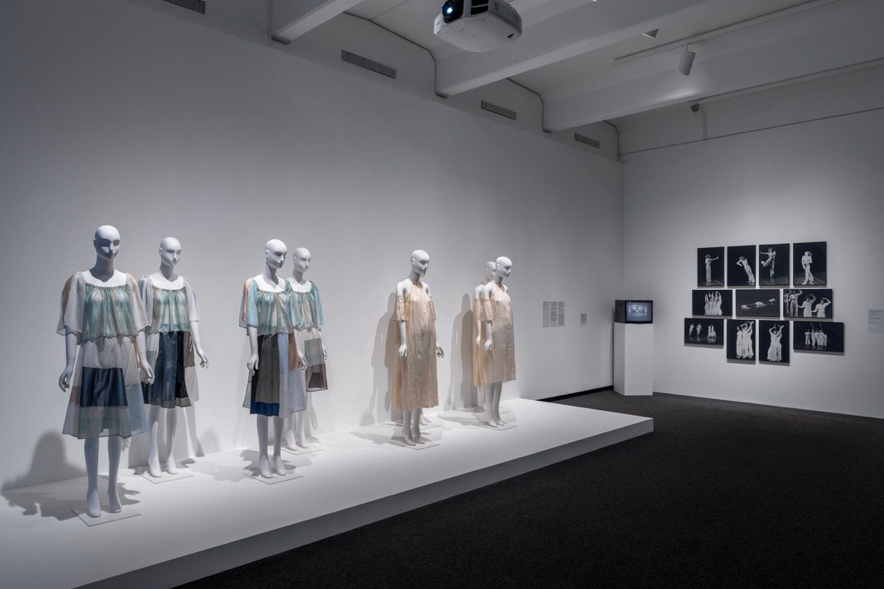 Installation view of a museum exhibition with mannequins in light-colored dresses on a platform. Black-and-white photographs and a video screen are displayed on the white wall in the background.