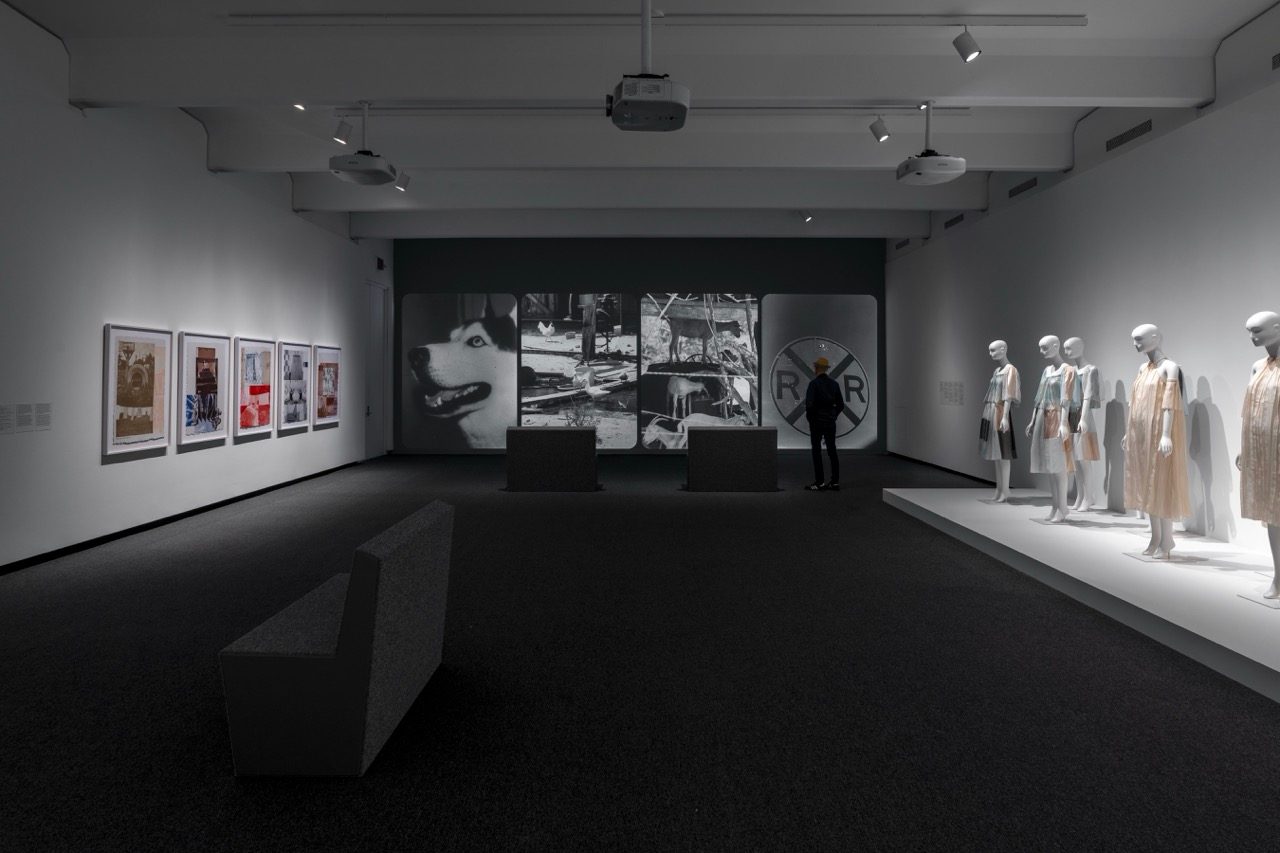 Installation view of a museum exhibition. Framed large-scale prints hang along the left wall, while several mannequins wearing light-colored garments stand on a raised platform along the right wall. Large black-and-white images including, at far right, a railroad crossing sign, are displayed at the back of the gallery, where a visitor stands observing.